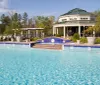 A serene outdoor swimming pool with a decorative fountain feature and a clubhouse in the background set against a backdrop of clear skies and greenery
