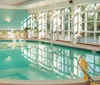 The image shows an indoor swimming pool with clear blue water surrounded by lounge chairs and large windows that allow ample natural light to fill the tranquil space