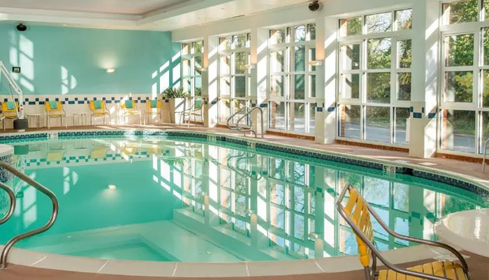 The image shows an indoor swimming pool with clear blue water surrounded by lounge chairs and large windows that allow ample natural light to fill the tranquil space