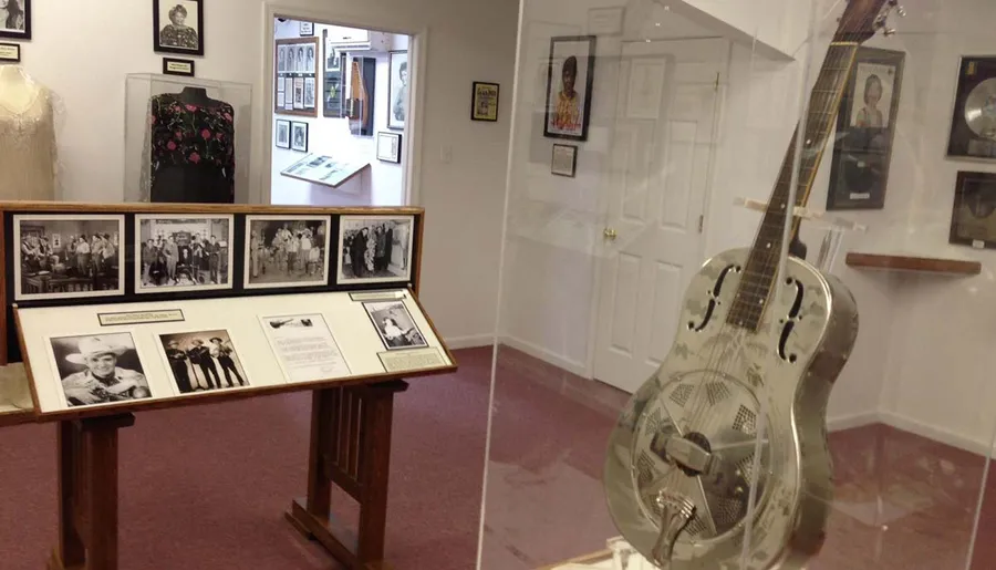 The image shows an interior view of a music-themed museum or gallery with framed photographs, artifacts, and a silver resonator guitar on display.