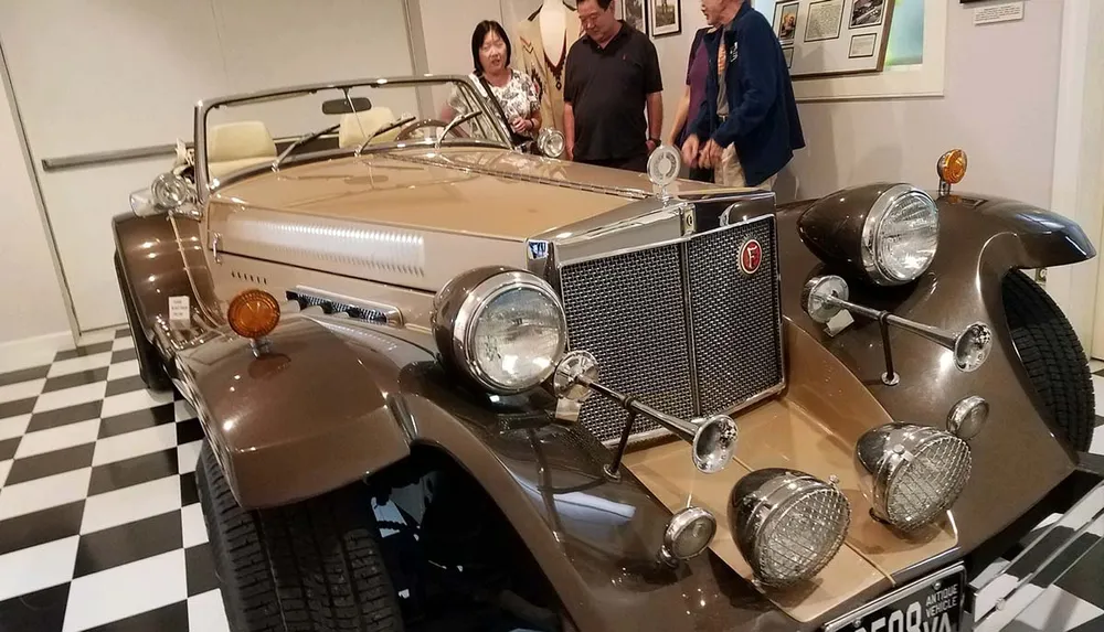 A group of people admires a classic tan convertible car with distinctive front grilles and multiple headlamps on display in what appears to be a museum with a black and white checkered floor