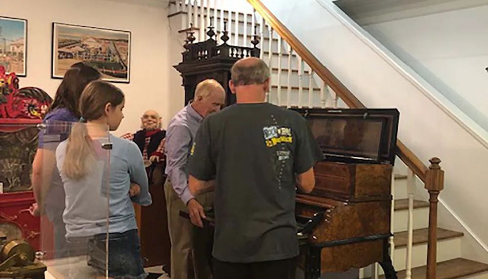A group of people is gathered around an antique piano in an indoor setting appearing to be engaged in a discussion or demonstration