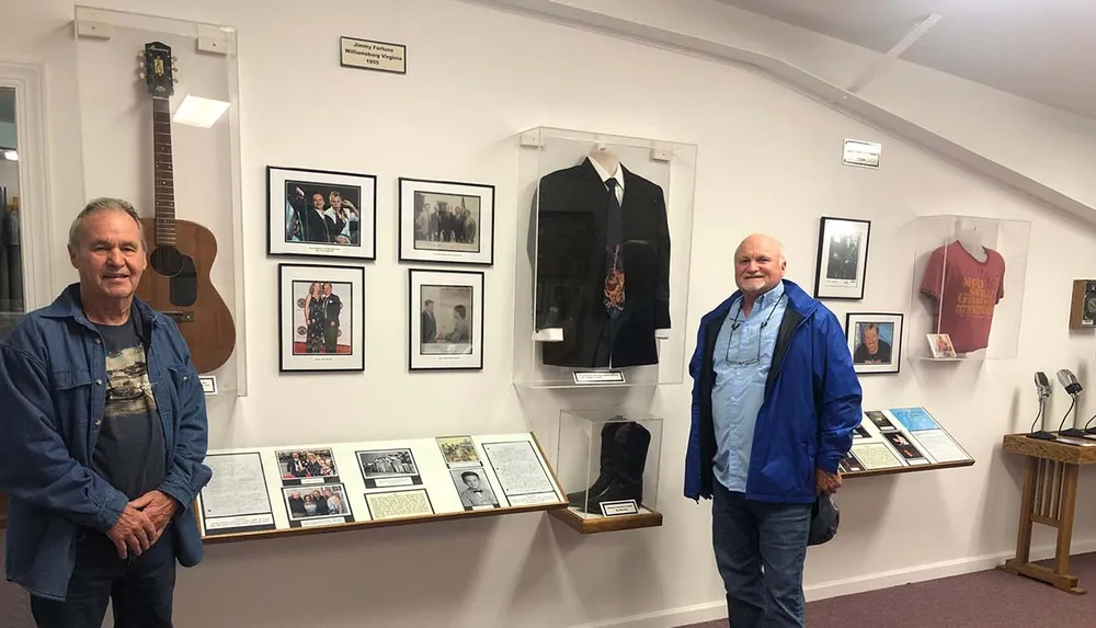 Two individuals are standing in a room that appears to be a museum or exhibit space surrounded by framed photographs memorabilia in display cases and information panels