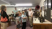 A group of people, including children and adults, are looking at exhibits inside a museum or gallery, with one man appearing to explain something to the group.