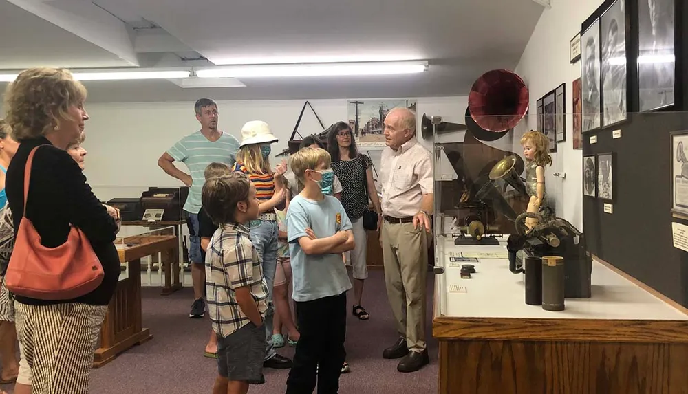 A group of people including children and adults are looking at exhibits inside a museum or gallery with one man appearing to explain something to the group