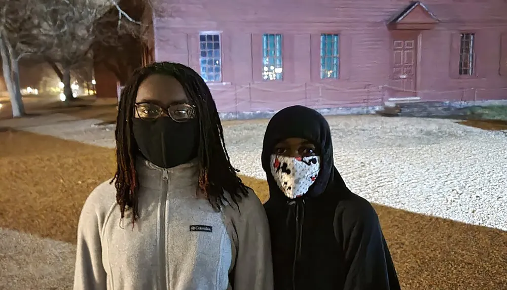 Two individuals are standing outdoors at night wearing masks with leafless trees and a pink building with lit windows in the background