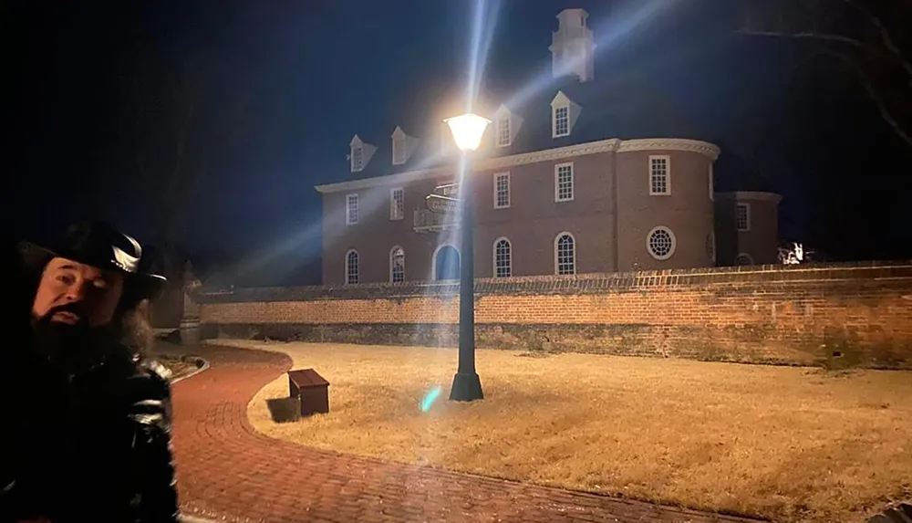 A person in historical costume stands to the left of the frame with an illuminated streetlamp and a colonial-style brick building in the background at night