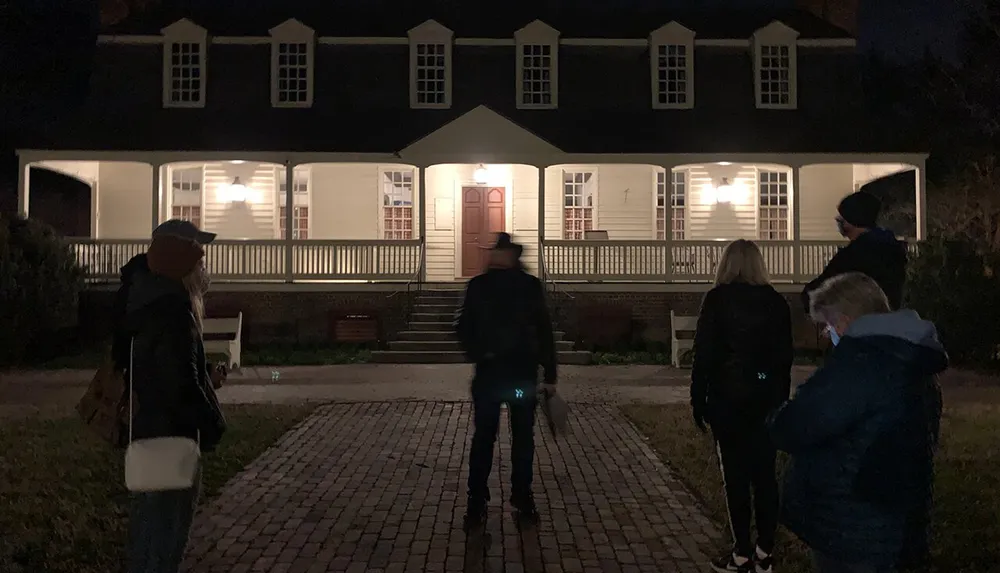 A group of people stands outside a well-lit traditional two-story house at night