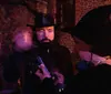 The image shows two individuals with one looking intently at a smartphone while the other wearing a pointed hat is turned away all against a backdrop of a dimly lit brick wall setting