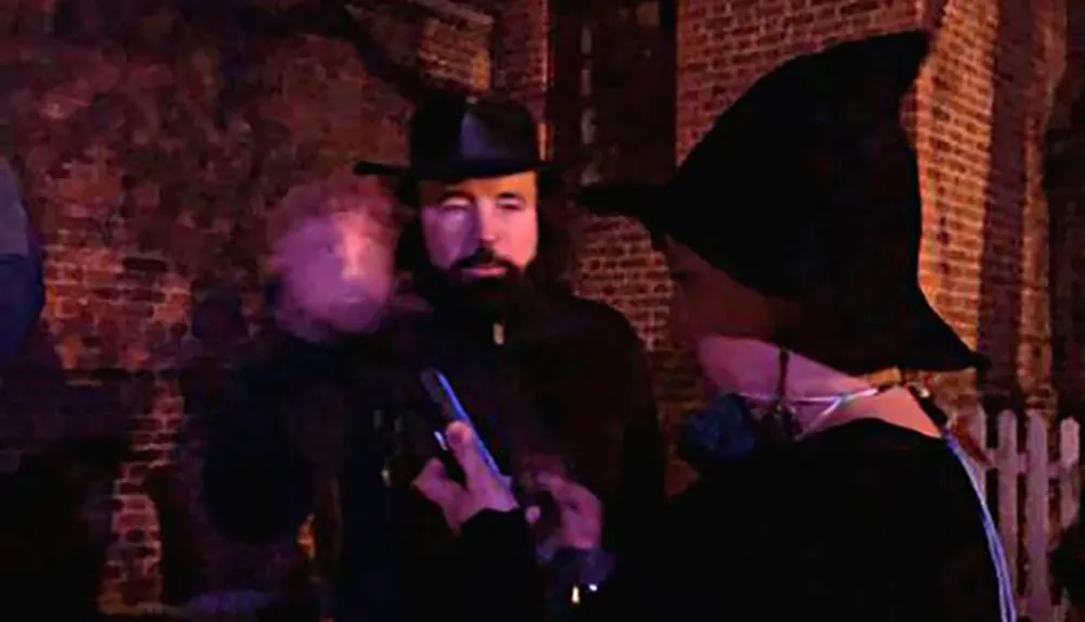 The image shows two individuals with one looking intently at a smartphone while the other wearing a pointed hat is turned away all against a backdrop of a dimly lit brick wall setting