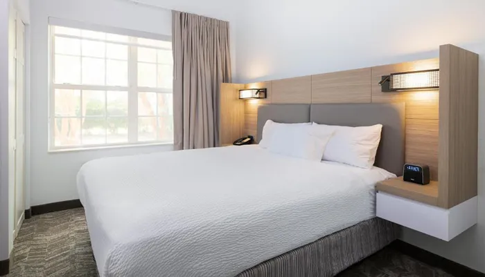 The image shows a neatly made bed with white bedding in a modern hotel room with a large window side tables and wall-mounted lamps