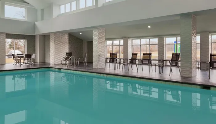 The image shows an indoor swimming pool with chairs along the side and large windows providing daylight