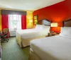 The image shows a bright and colorful hotel room with two beds a red accent wall and a view out the window