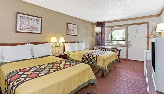 The image shows a modestly furnished hotel room with two queen-sized beds a patterned carpet and a small dining area by the window