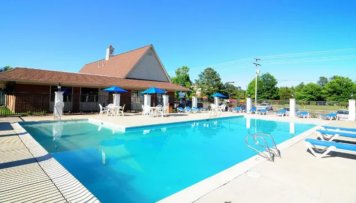 The image shows a serene outdoor swimming pool area with blue umbrellas lounge chairs and a building in the background under a clear blue sky