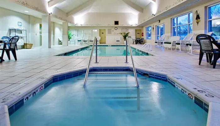This is an indoor swimming pool area with a jacuzzi multiple lounge chairs and large windows allowing in natural light