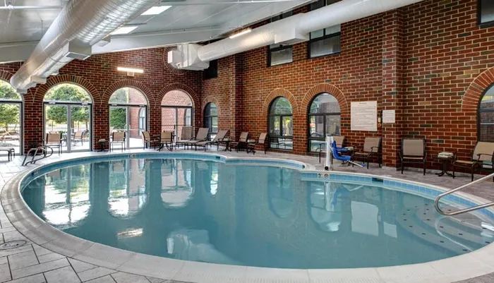 An indoor swimming pool with a curved design is surrounded by multiple chairs and features large windows within a brick-walled space