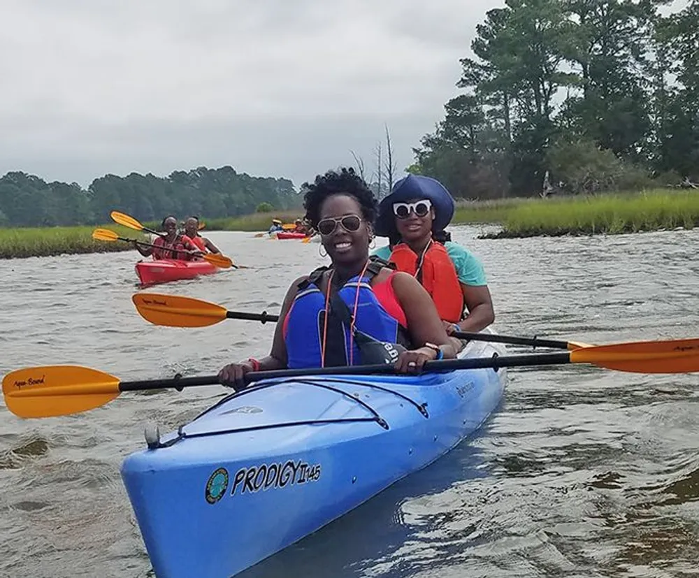 A group of people are kayaking in a calm waterway with two smiling individuals in the foreground paddling a blue tandem kayak