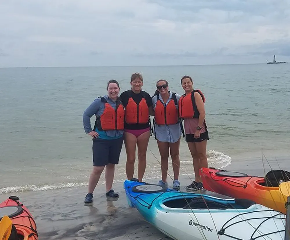 Four individuals wearing life jackets pose on a beach with kayaks suggesting they are about to embark on or have returned from a kayaking adventure