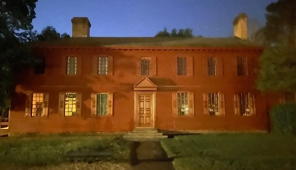 The image shows a two-story red-painted building with glowing windows under a dusk or night sky creating a warm and somewhat eerie atmosphere