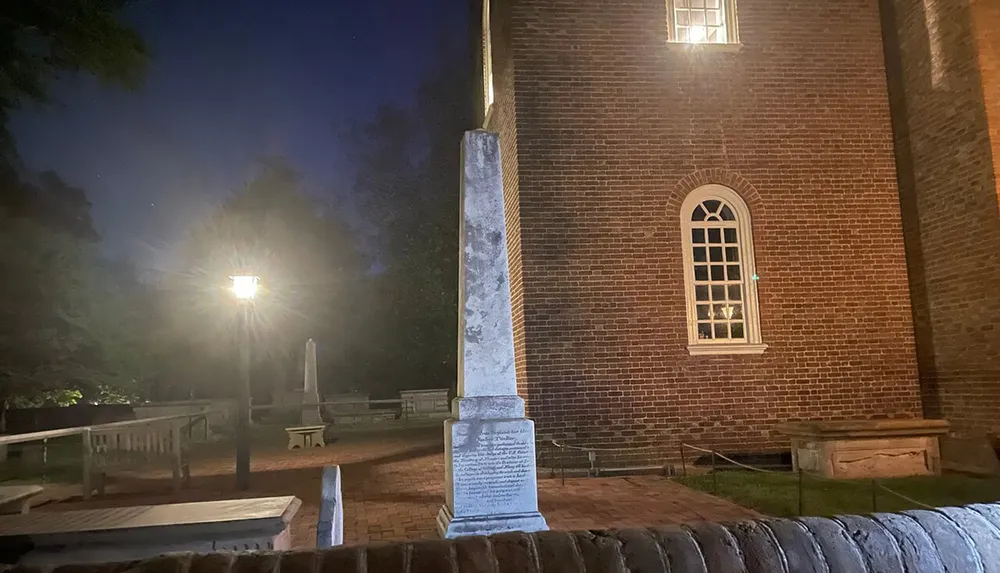 The image shows an illuminated historical brick building at night with an obelisk monument in the foreground likely located within a cemetery or memorial park