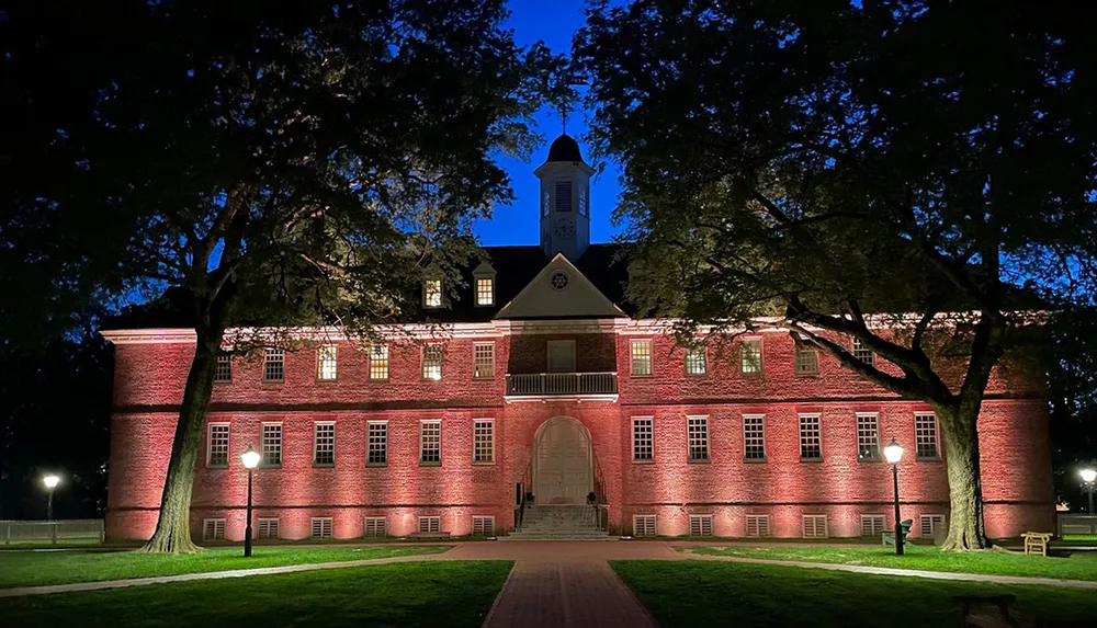 A colonial-era brick building illuminated at night by warm lights against a dusky blue sky framed by silhouettes of trees