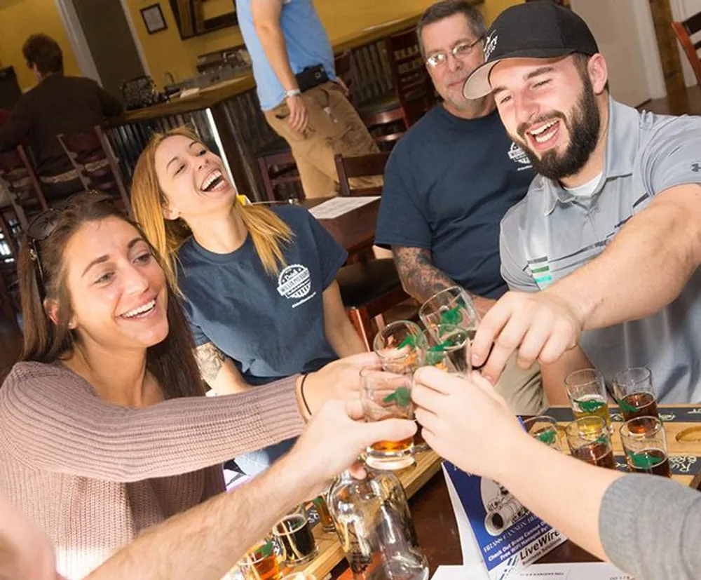 A group of cheerful people are toasting with glasses at a table in what looks like a pub or brewery