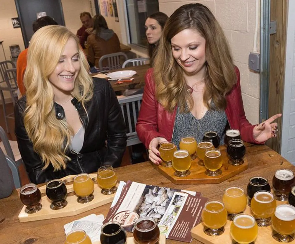 Two women are smiling and looking at a selection of different beers arranged on a paddle for tasting in a casual indoor setting