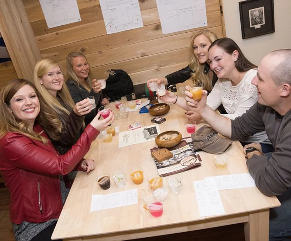 A group of friends are toasting with drinks around a small table filled with various beverages and snacks