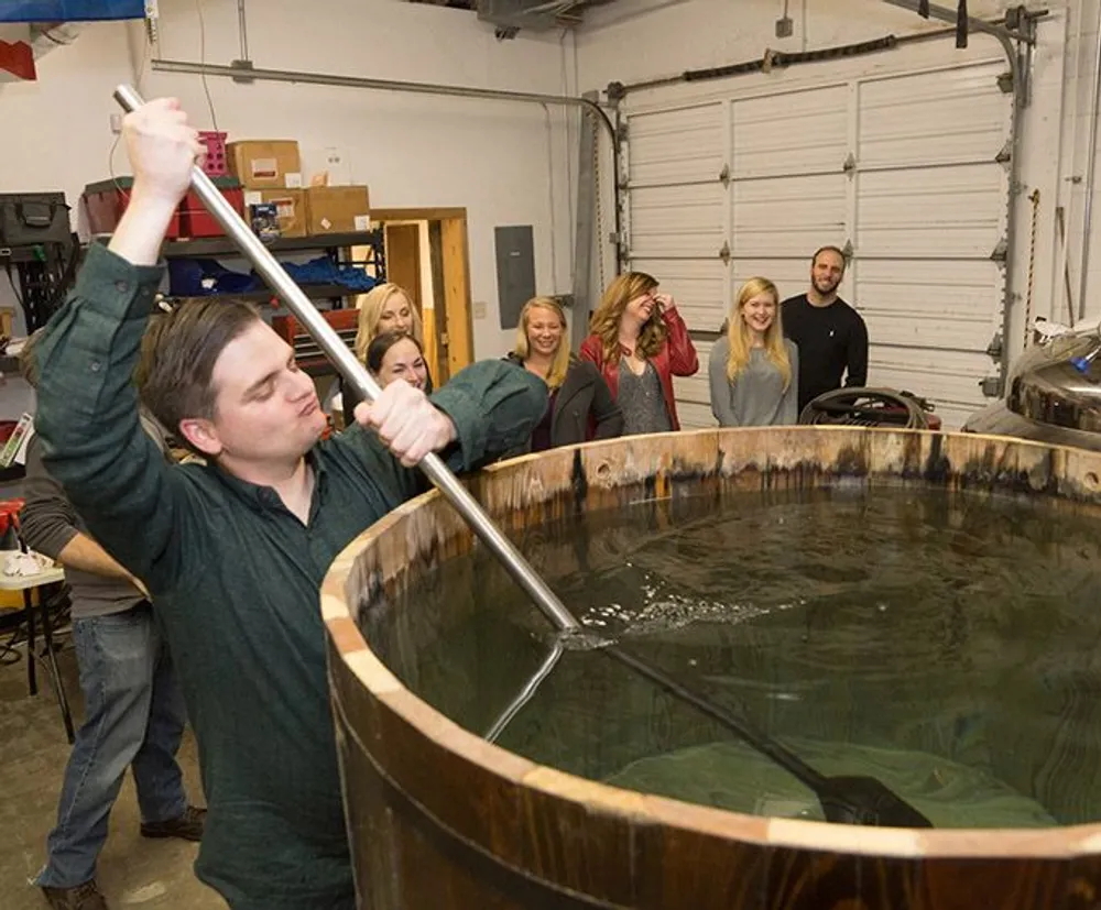 A person is stirring a large wooden vat with a long pole while several onlookers watch some of whom are smiling or laughing