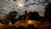 A house is illuminated by warm lights at night under a cloudy, moonlit sky.
