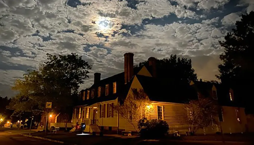 A house is illuminated by warm lights at night under a cloudy moonlit sky