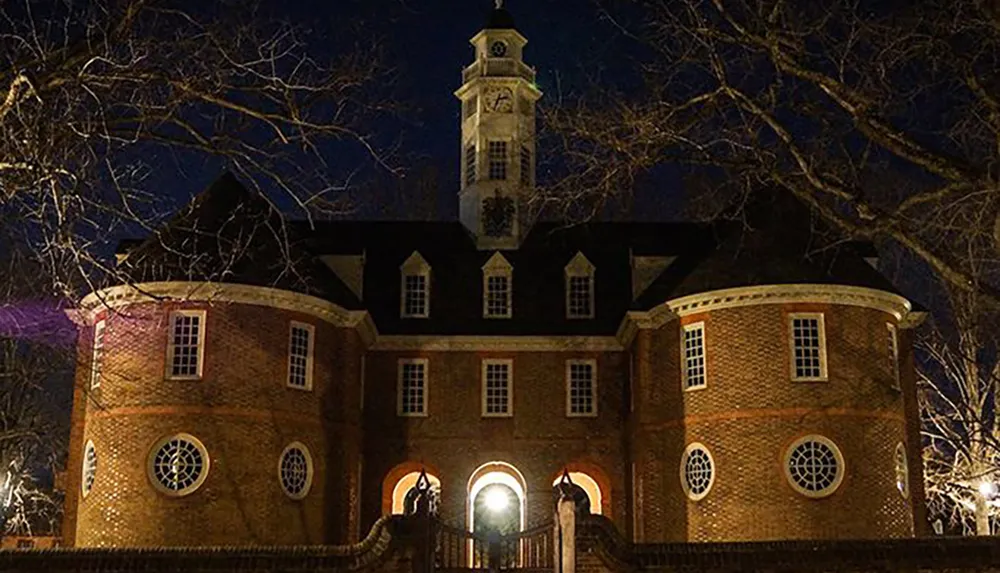 A historic brick building with a central tower is illuminated at night framed by bare tree branches