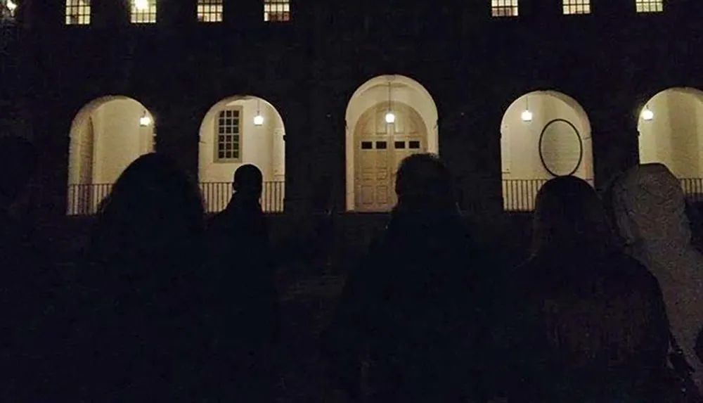 A group of people is standing in front of a large building with arched doorways and lit windows at night