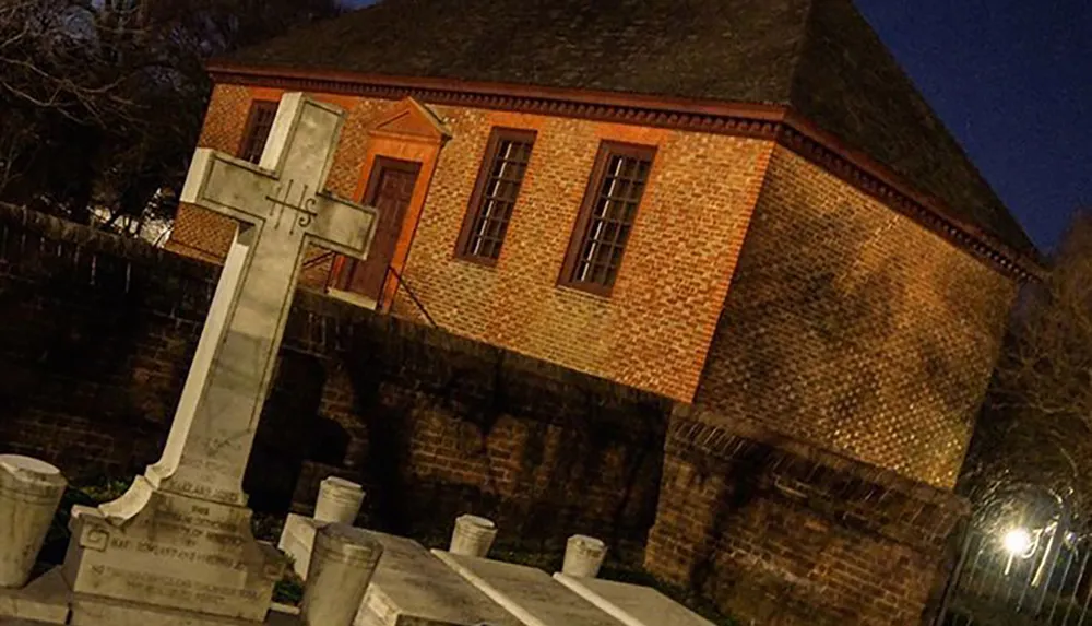 The image shows a gravestone with a cross in the foreground against the backdrop of a brick building at night
