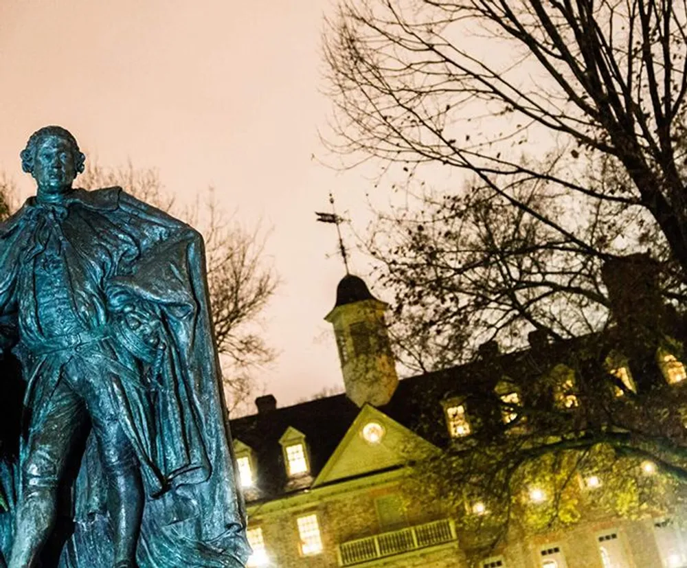 An illuminated bronze statue stands in the foreground with a historic building and a leafless tree in the background against a dusky sky