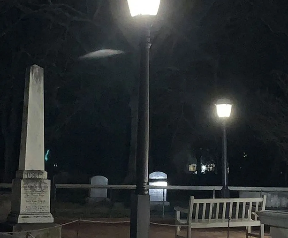 The image depicts a dimly lit outdoor scene at night with street lamps illuminating a park or cemetery area containing benches a monument and bare trees in the background