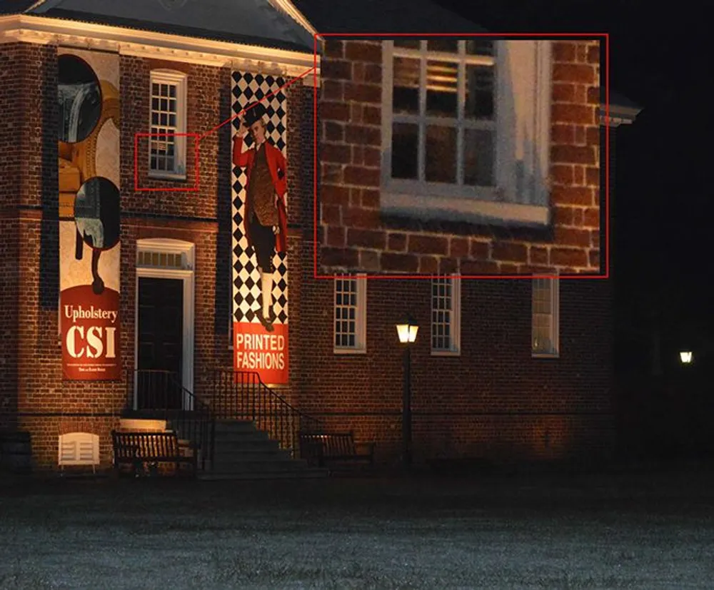 This image shows a nighttime view of a brick building with illuminated windows and posters on the exterior wall with one window zoomed in on in a red inset possibly suggesting something of interest there