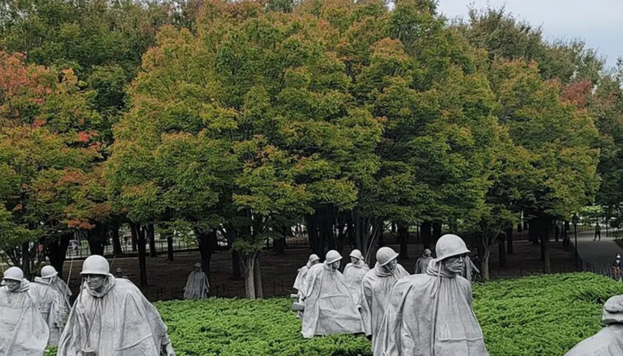 Statues depicting a group of soldiers stand amidst greenery with a backdrop of trees showing early autumn colors.