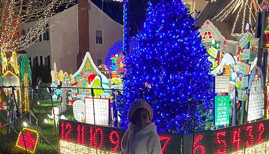 A person stands in front of a house extravagantly decorated with vibrant Christmas lights and festive displays.