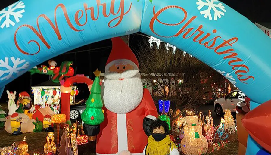 A vibrant outdoor Christmas display featuring an inflatable Santa Claus, a colorful arch reading Merry Christmas, and various illuminated decorations, with a person standing in front.