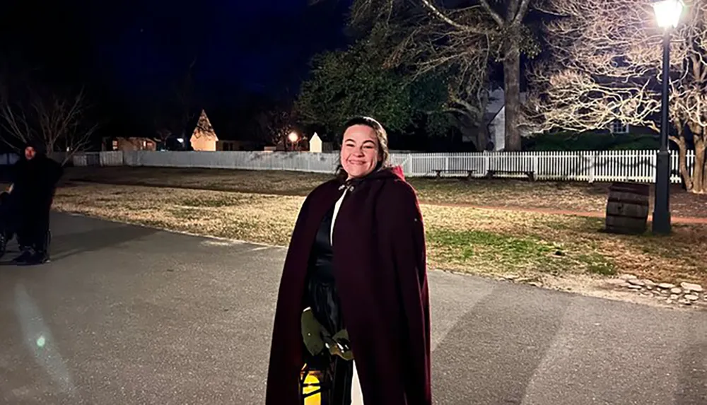 A smiling person wearing a cape stands in an outdoor nighttime setting with a well-lit path trees and a historic-looking building in the background