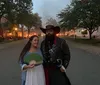 A smiling couple is posing for a photo at twilight with a historic brick building in the background dressed in what appears to be period costumes