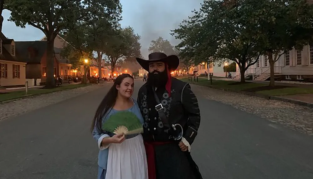 A woman holding a fan stands next to a man dressed in historical costume on an old-fashioned street at dusk