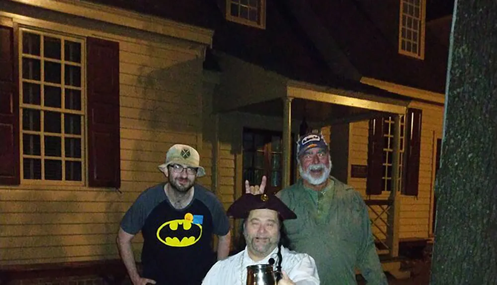 Three individuals are posing for a photo at night in front of a house with one person playfully giving bunny ears to another