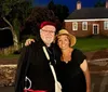 A smiling couple is posing for a photo at twilight with a historic brick building in the background dressed in what appears to be period costumes