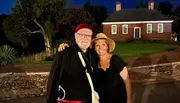 A smiling couple is posing for a photo at twilight with a historic brick building in the background, dressed in what appears to be period costumes.