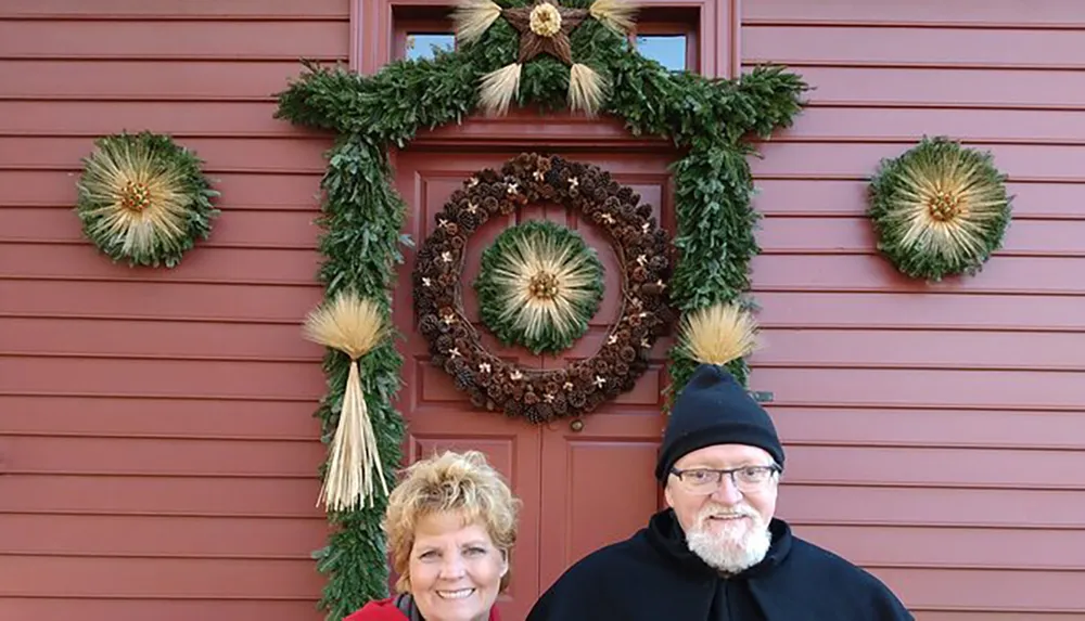 A smiling couple is posing in front of a red door decorated with a festive Christmas garland and a wreath
