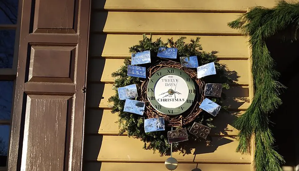 A Christmas-themed wreath with a clock face reading The Twelve Days of Christmas is hung on a yellow door decorated with greenery and additional images portraying winter or holiday scenes