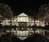 The image shows a large stately house beautifully illuminated with white holiday lights reflected in a calm body of water in front of it